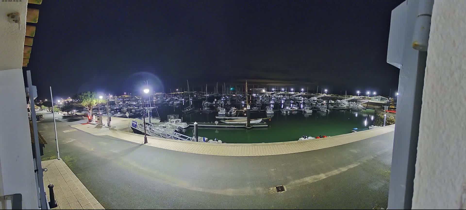 A marina filled with boats is seen at night under a clear, dark sky, with a paved area and streetlights visible.