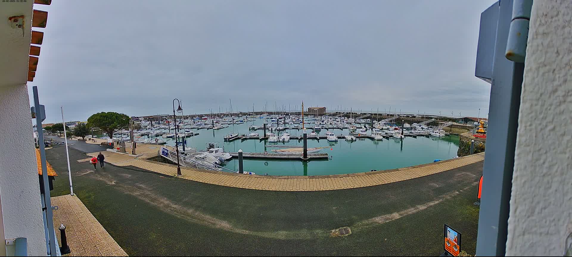 A marina filled with numerous boats is seen under an overcast sky, with two people walking along a paved path nearby.