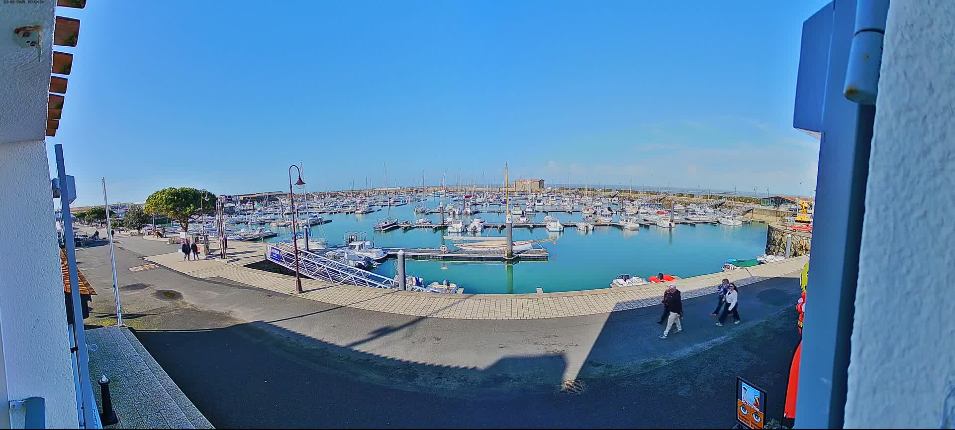 A marina filled with numerous boats is seen on a sunny day, with a few people walking along the paved walkway next to the water.
