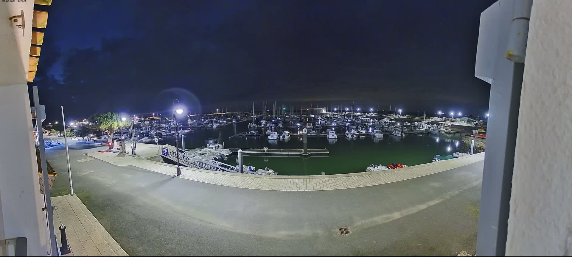 A nighttime view of a marina filled with numerous boats, illuminated by artificial lights under a dark, partly cloudy sky.