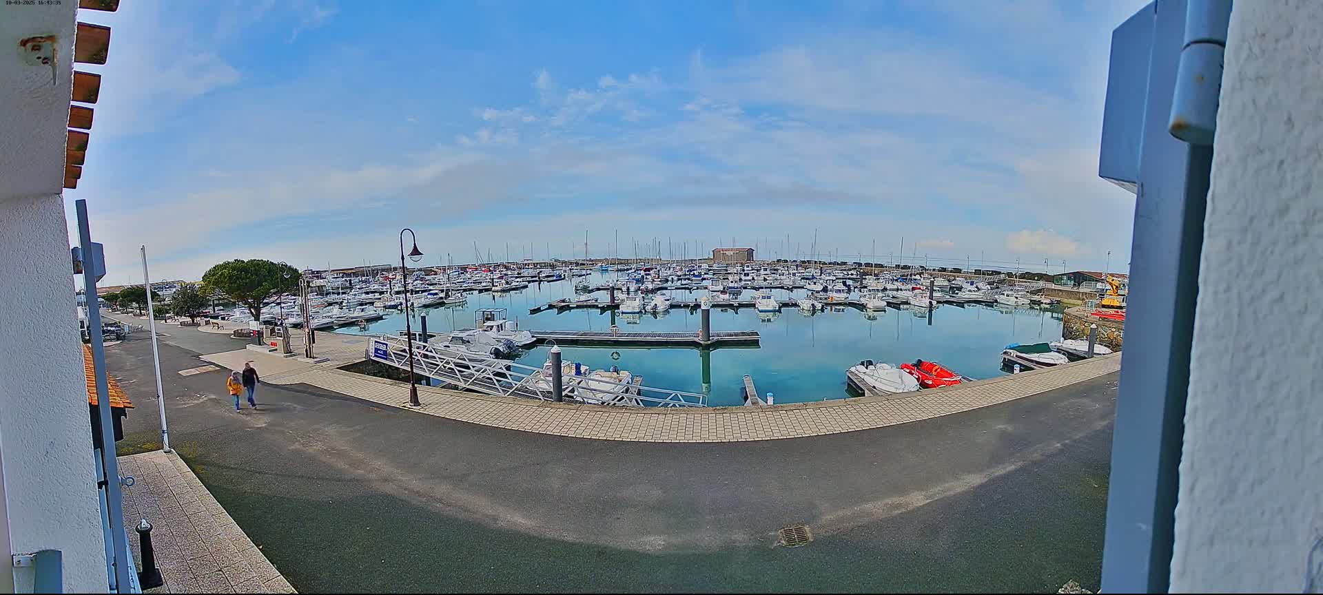 A marina filled with numerous boats on a calm, sunny day, with two people walking along a paved path nearby.
