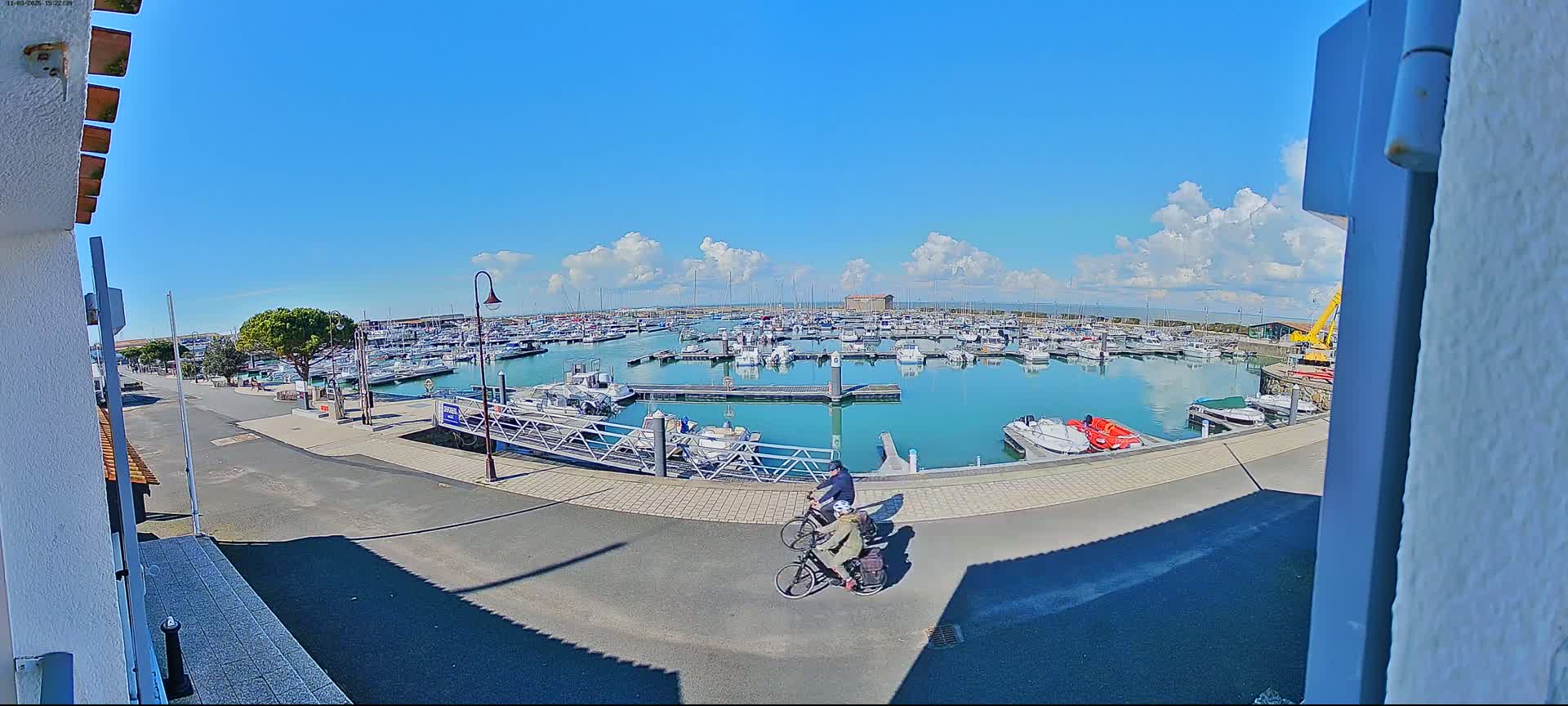 Two people ride bicycles along a paved path overlooking a marina filled with boats under a partly cloudy blue sky.