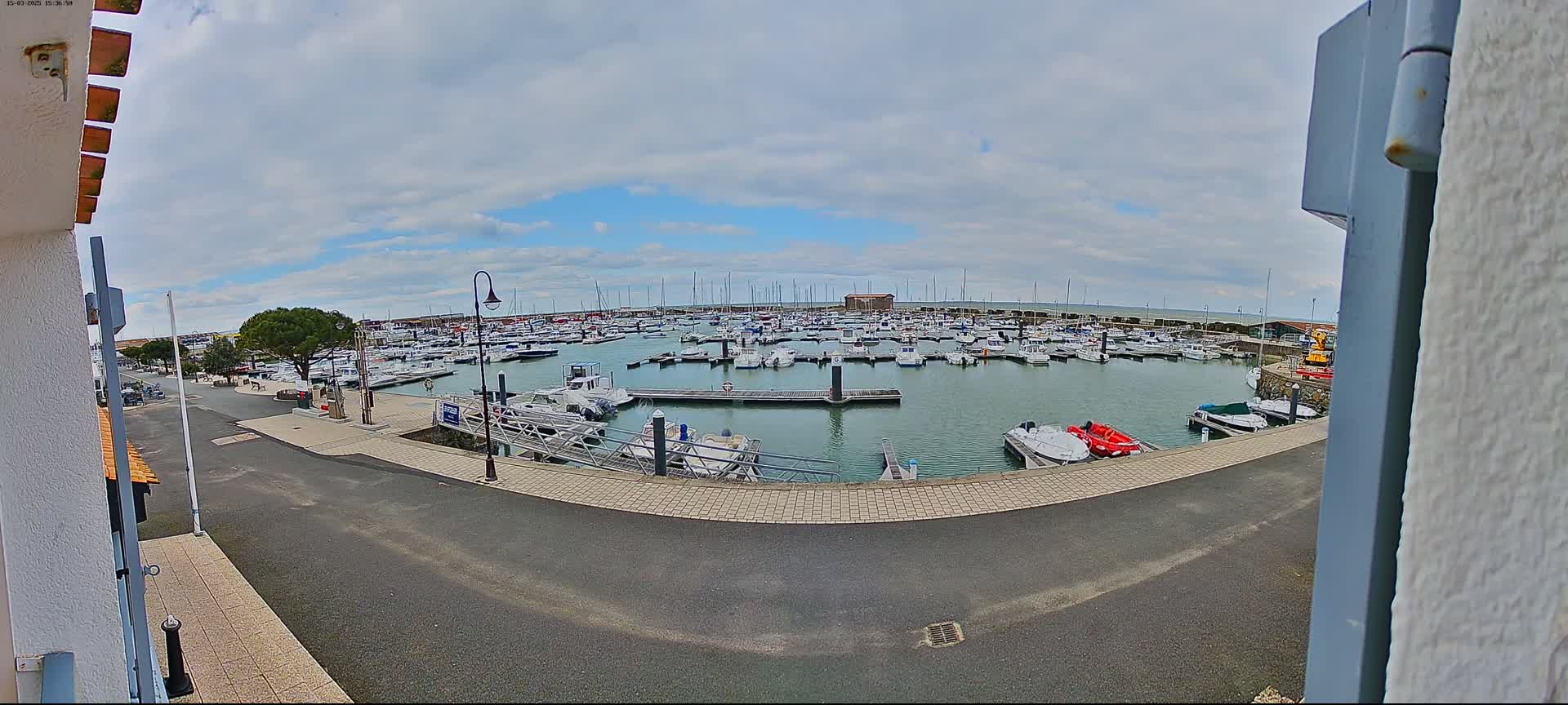 A marina filled with numerous boats is viewed from a paved area on a partly cloudy day.