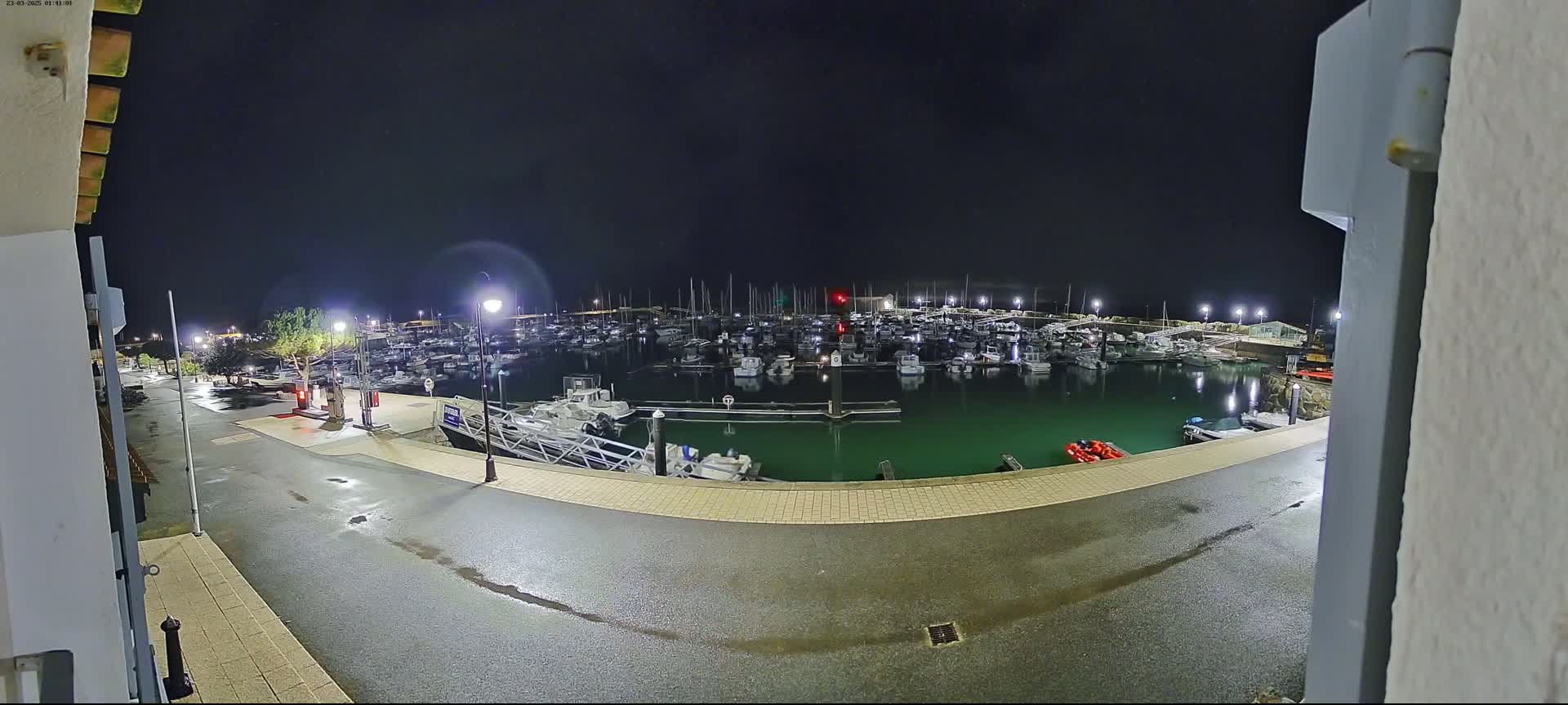 A marina filled with numerous boats is seen at night under a clear, dark sky, with a wet paved area visible in the foreground.
