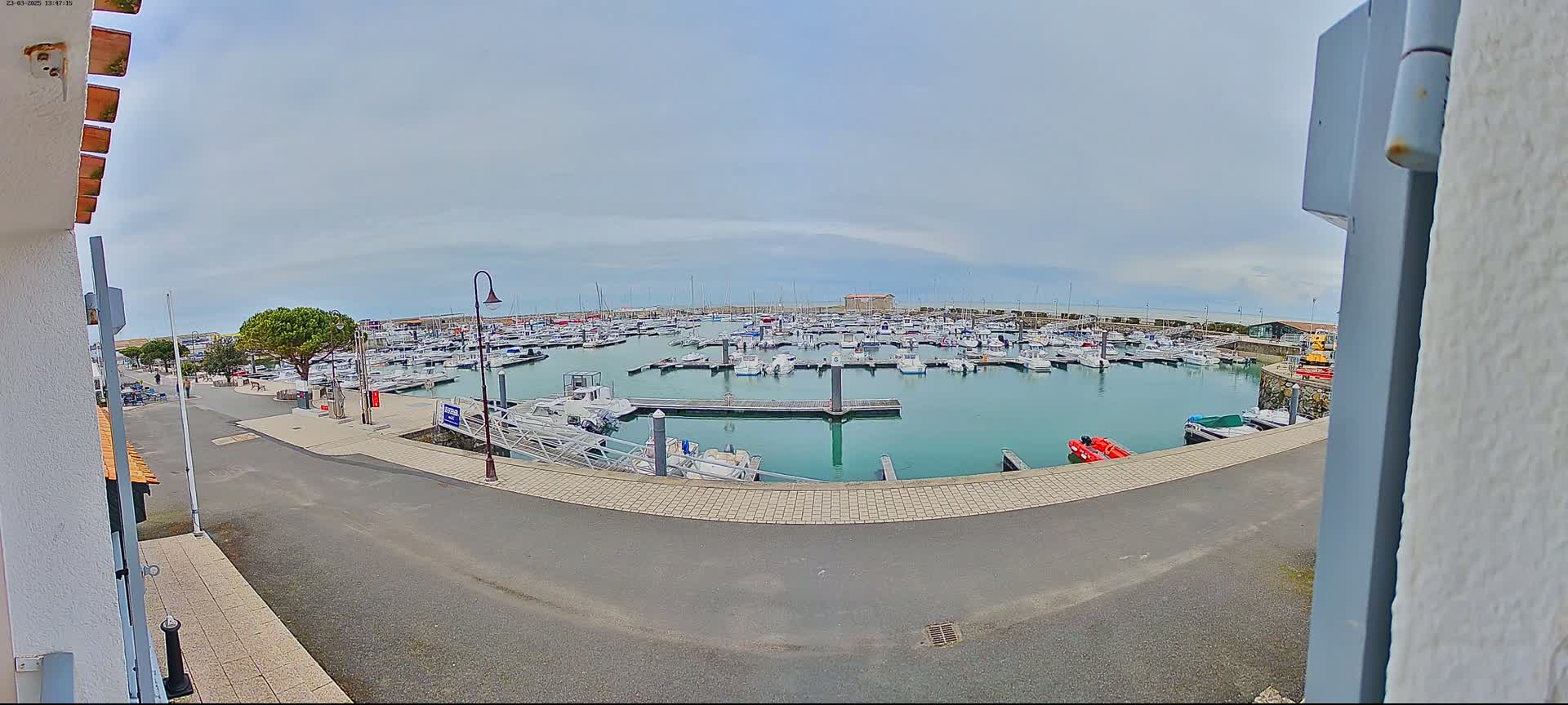 A marina filled with numerous boats sits on calm, greenish-blue water under a mostly cloudy sky.