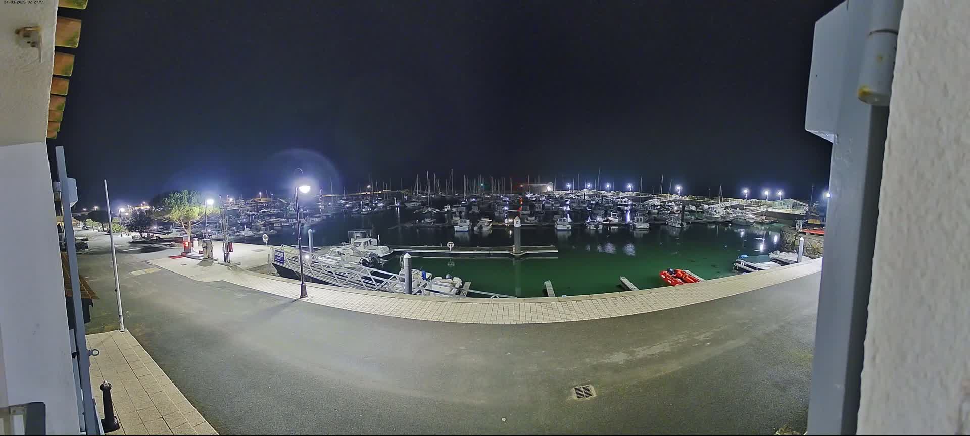 At night, many boats are docked in a marina, illuminated by lights, with a paved area visible in the foreground.