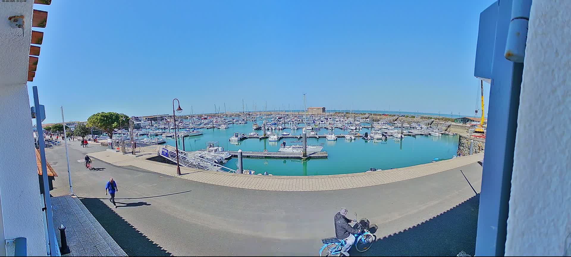 A marina filled with numerous boats is seen on a sunny day, with a few people walking and cycling along a nearby roadway.