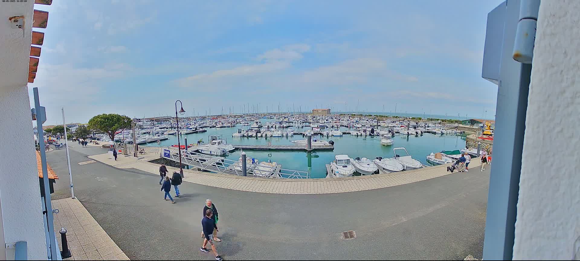 A marina filled with numerous boats on a sunny day, with several people walking along the paved area adjacent to the water.