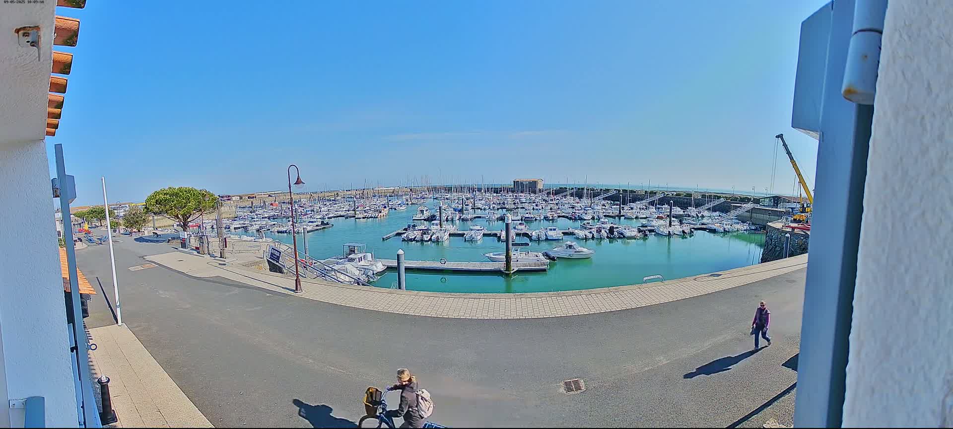 A marina filled with numerous boats is seen on a sunny day from a slightly elevated vantage point, with a couple of pedestrians walking along a nearby road.