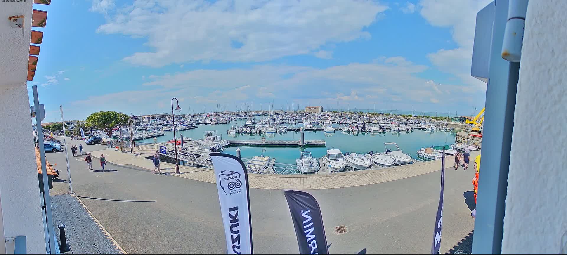 A marina filled with numerous boats is seen on a partly sunny day, with people walking along the paved waterfront.