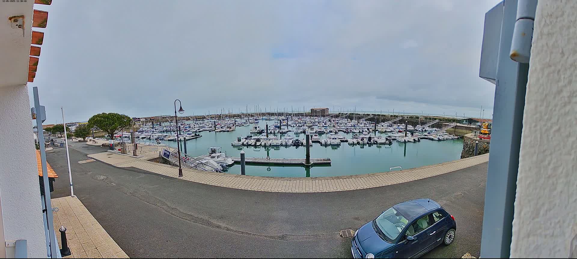 A marina filled with numerous boats is seen under an overcast sky, with a dark-blue car parked nearby.