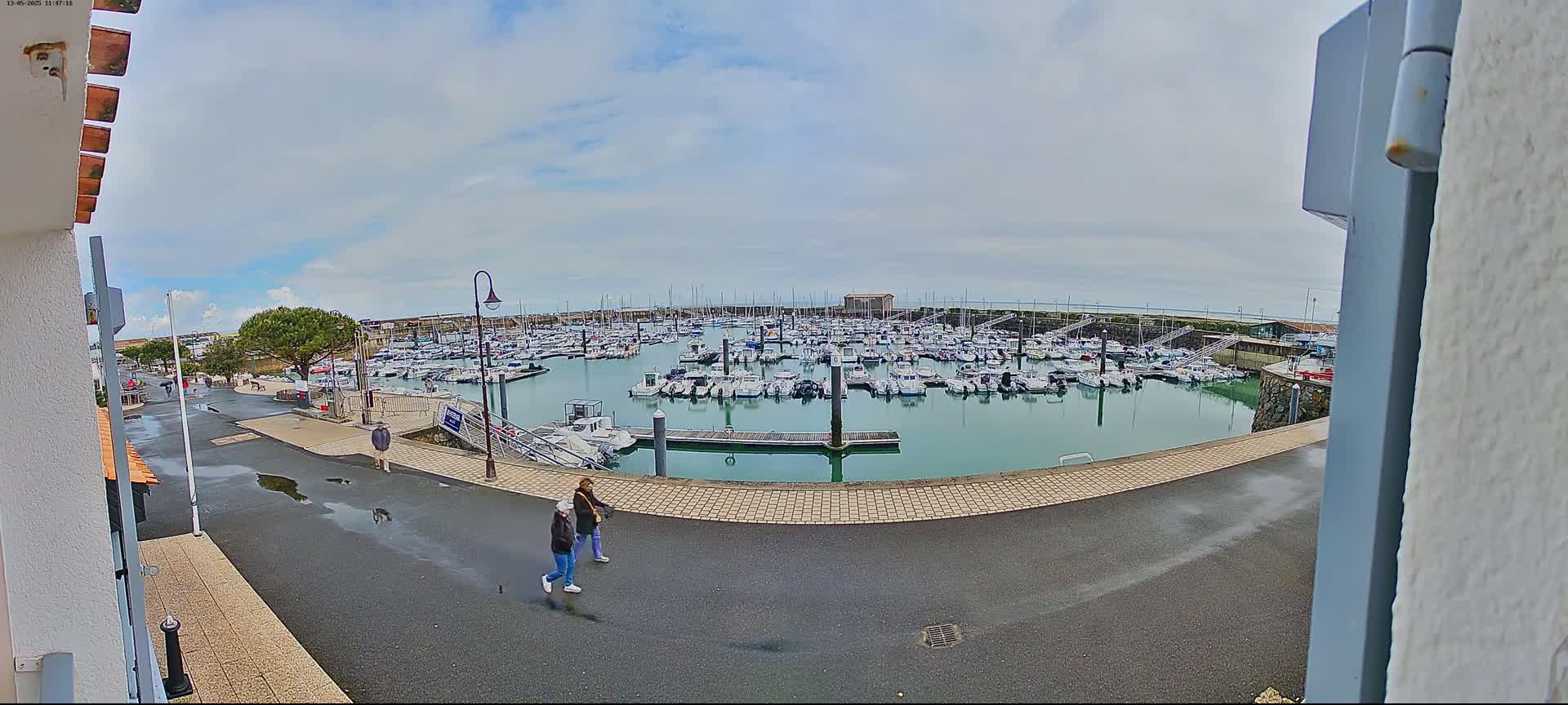 Two people walk along a paved path overlooking a calm harbor filled with numerous boats under a partly cloudy sky.
