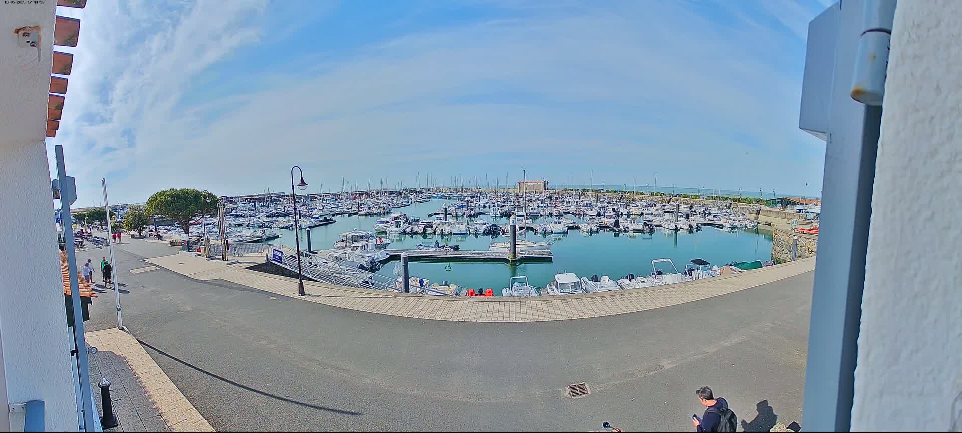 A marina filled with numerous boats is seen on a sunny day, with a few people walking along the paved area nearby.