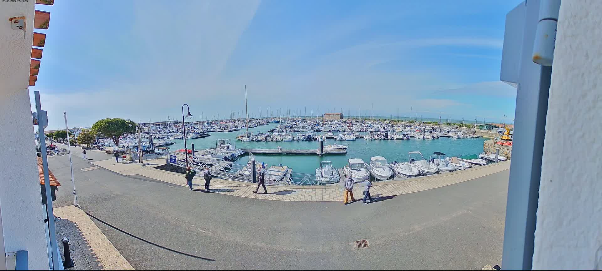 A marina filled with numerous boats is seen on a sunny day, with a few people walking along the paved area beside the water.