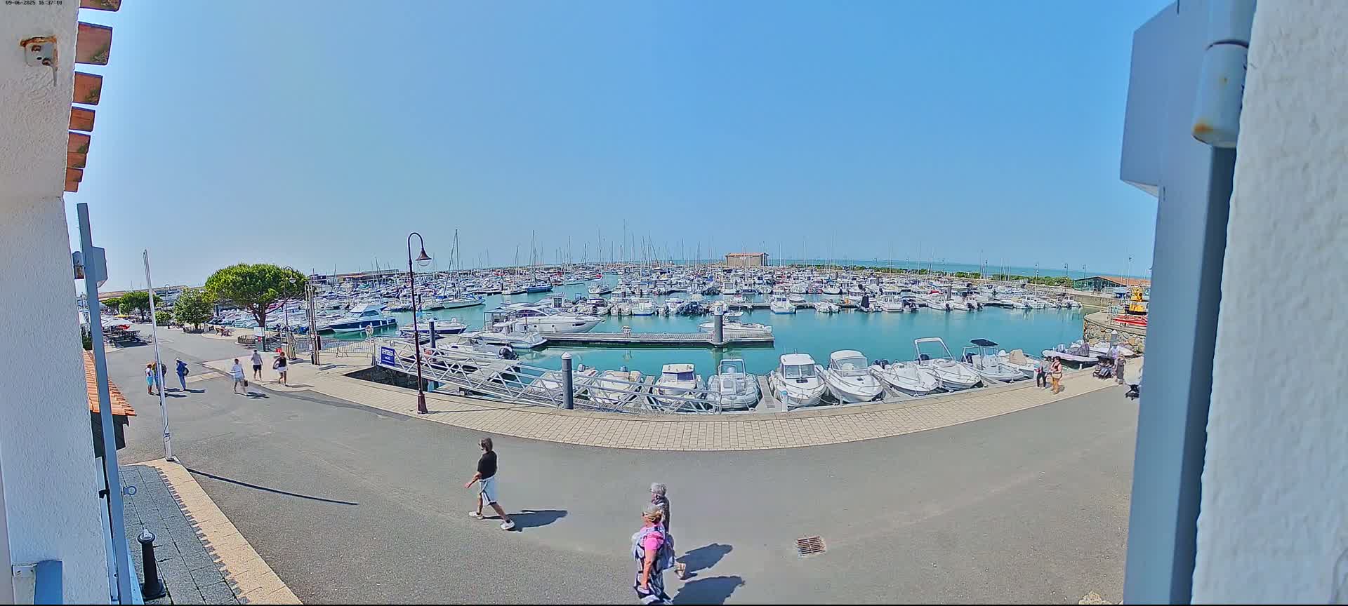 A marina filled with many boats is seen on a sunny day, with a few people walking along a paved path nearby.