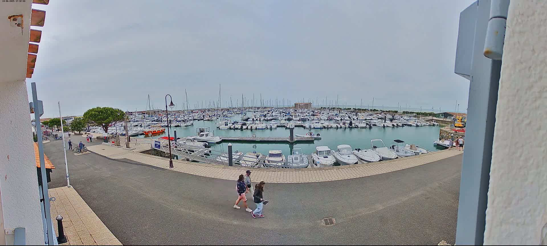 A marina filled with numerous boats under an overcast sky, with three people walking along the paved area next to the water.