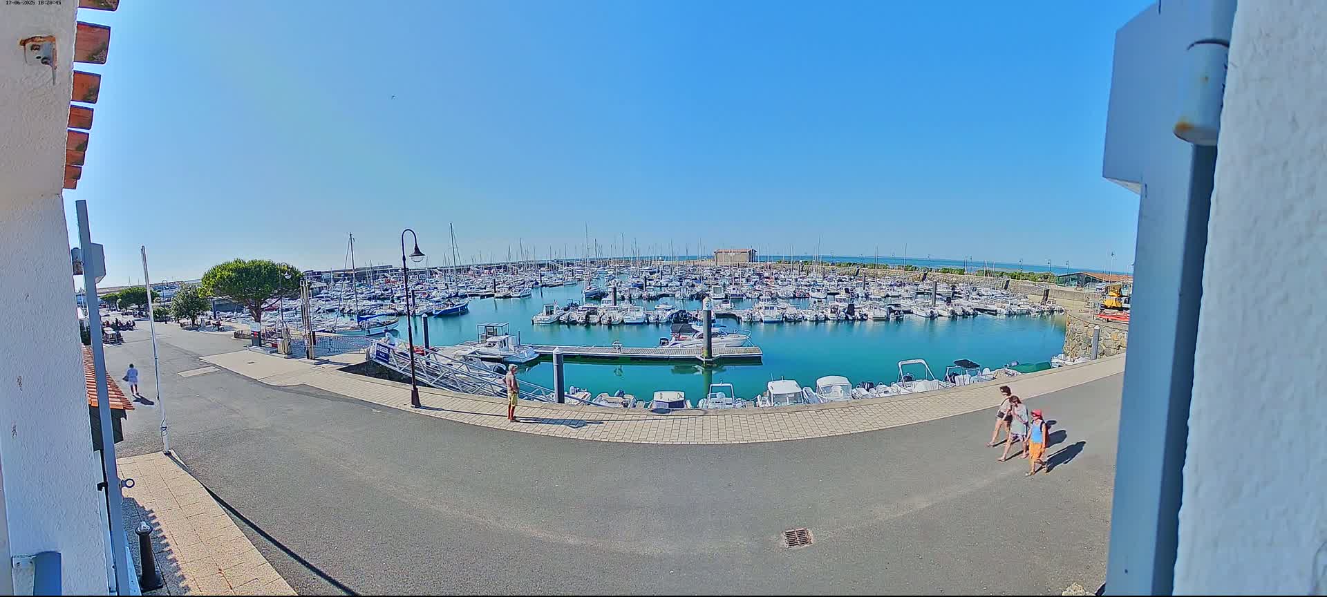 A marina filled with numerous boats is seen on a sunny day, with a few people walking along the paved area next to the water.