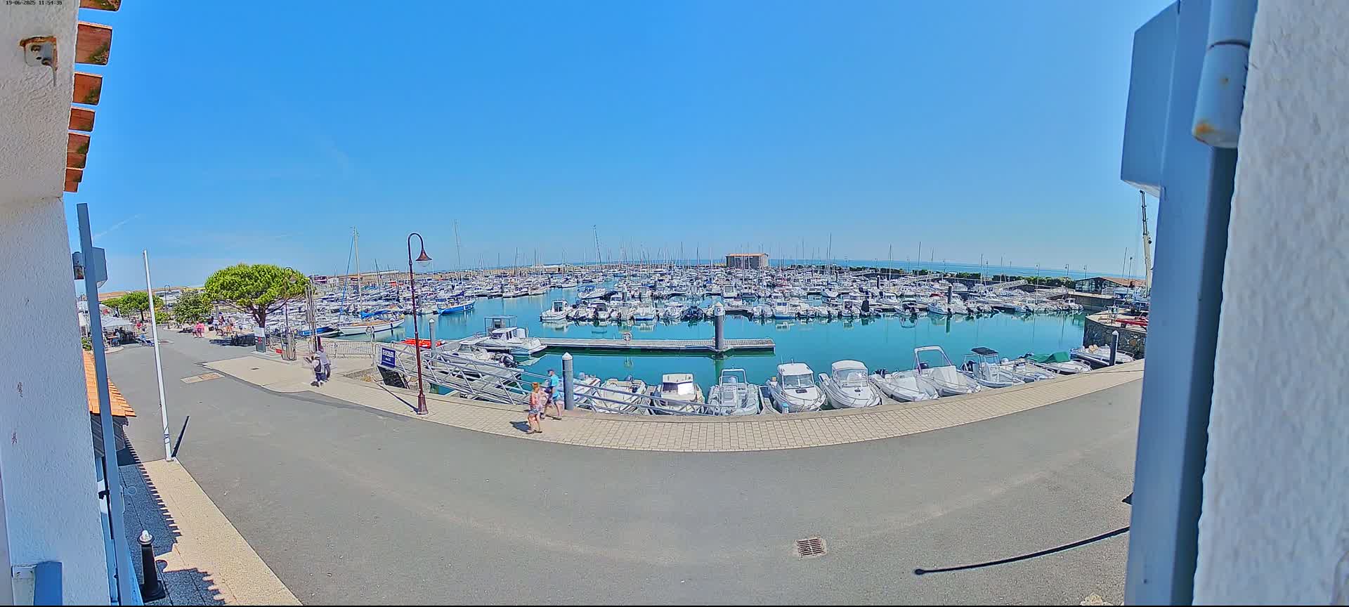 A marina filled with numerous boats is seen on a sunny day, with a few people walking along a paved path nearby.