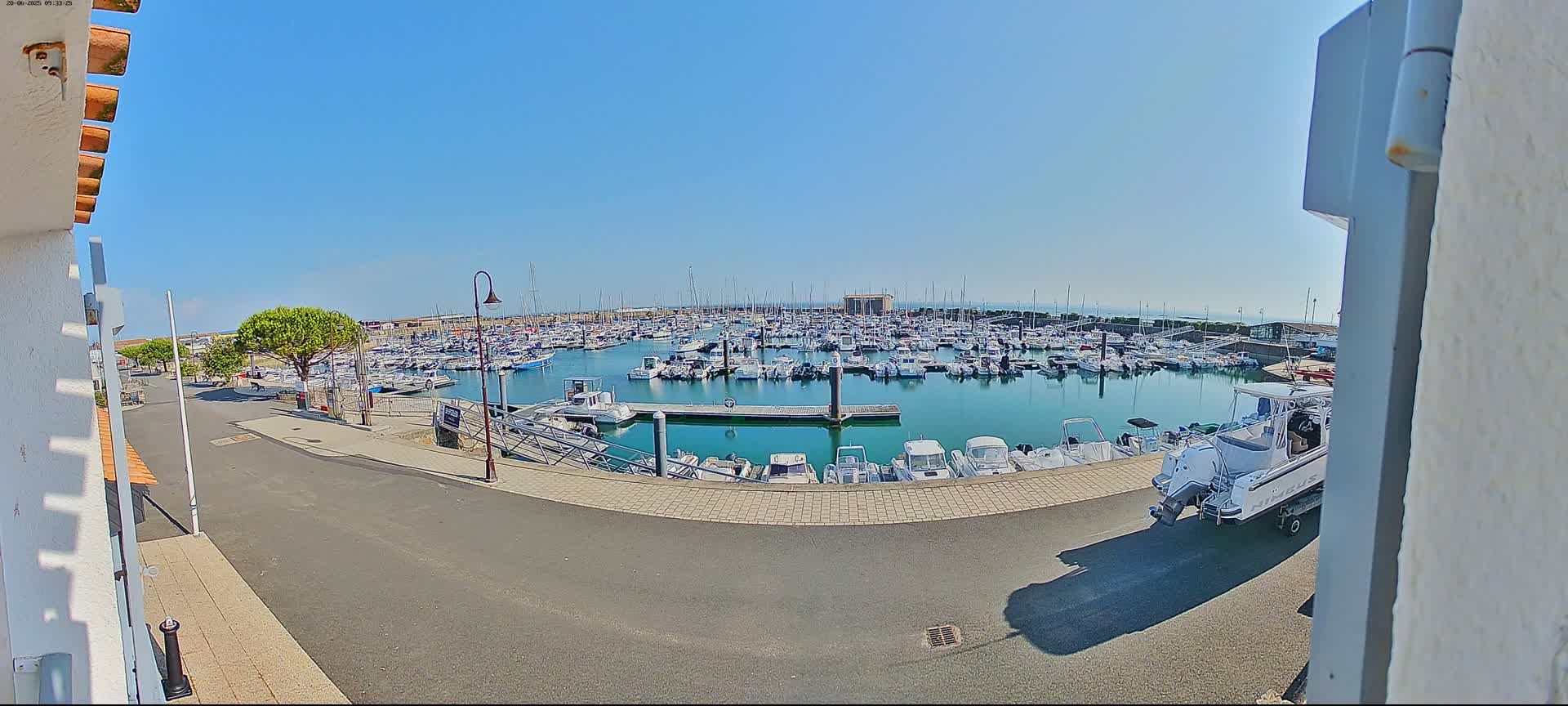 A sunny day shows a harbor filled with numerous boats, and a single boat on a trailer is parked on the road next to the harbor.