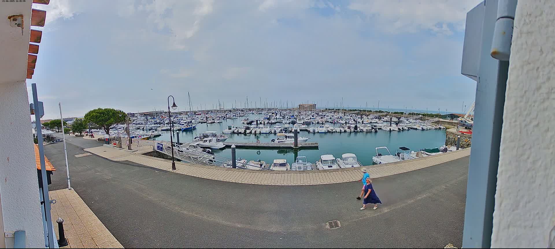 A couple walks along a paved path overlooking a marina filled with numerous boats under a partly cloudy sky.