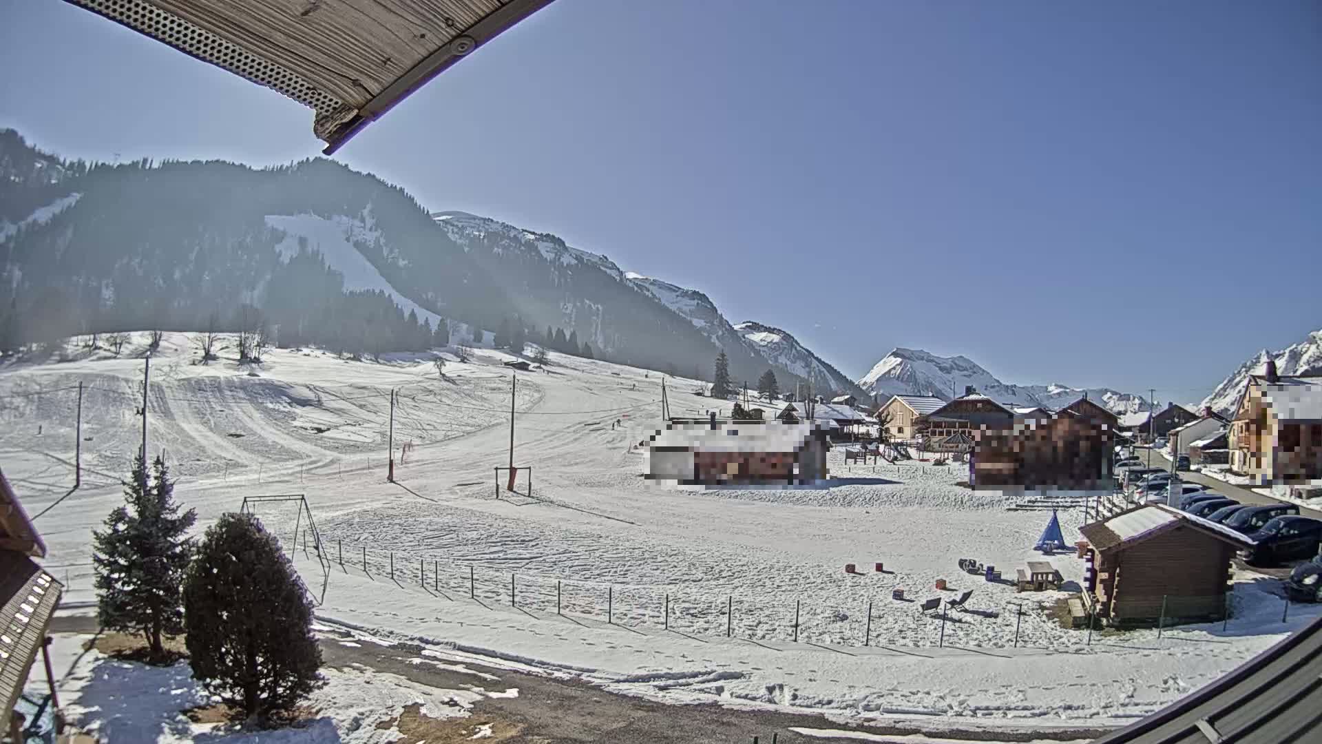 A snow-covered landscape with small buildings and a ski slope is visible under a clear, sunny sky, nestled against a backdrop of mountains.