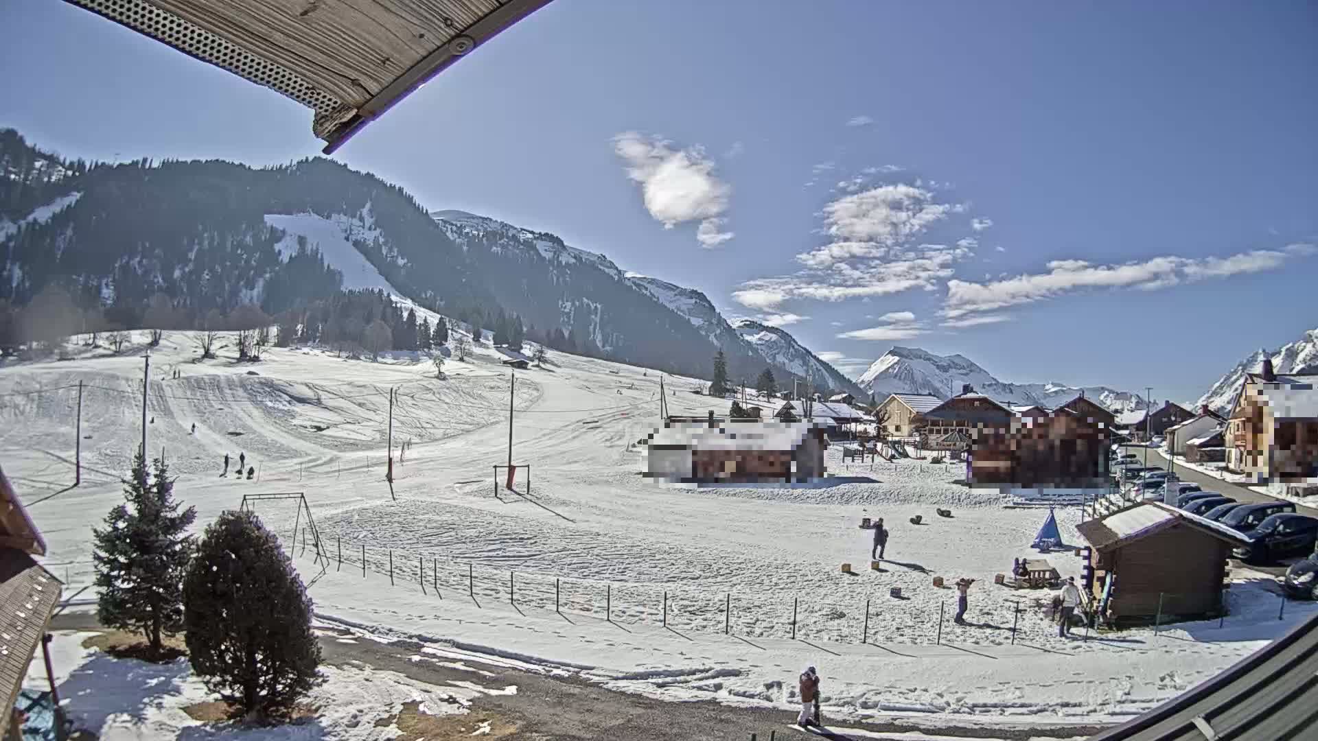 A snow-covered village nestled in a valley between mountains on a partly sunny day, with several people visible in the snow.