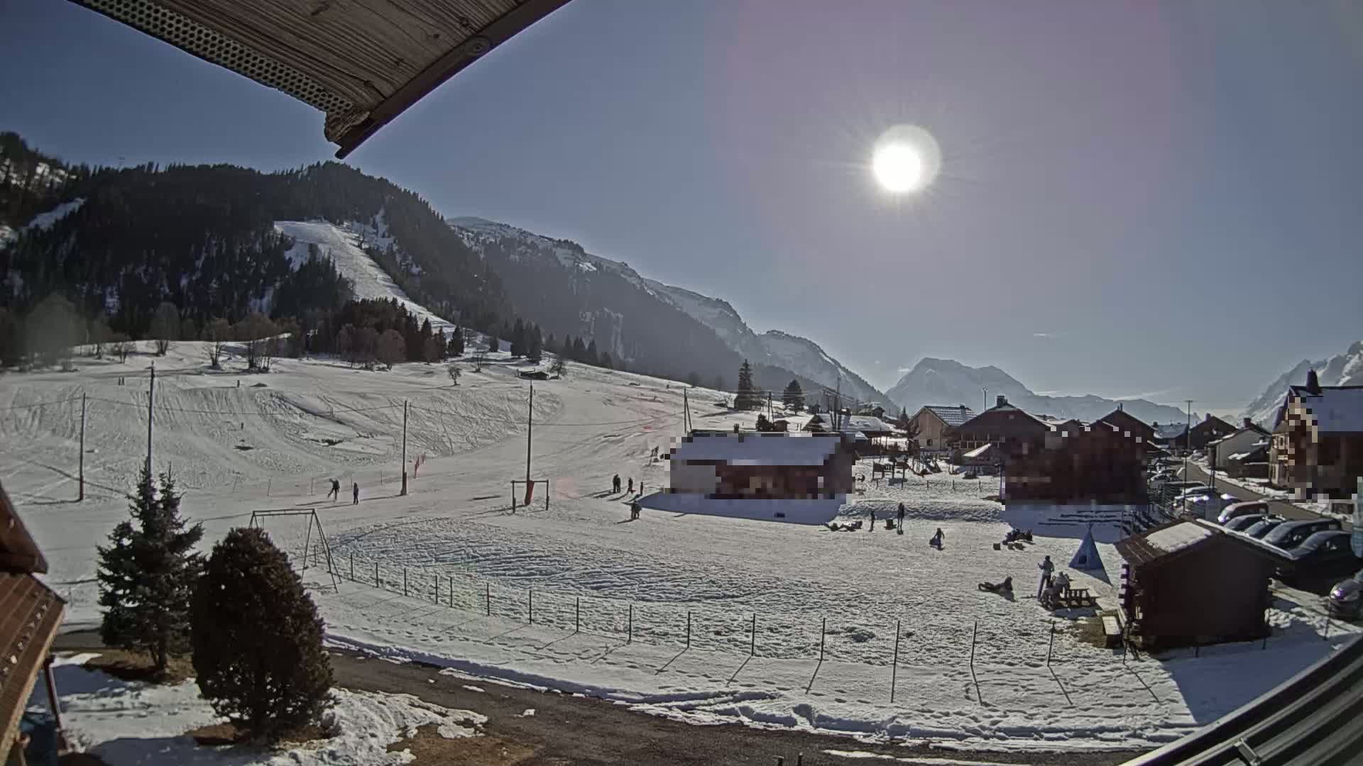 A sunny day in a snow-covered mountain village shows a small ski slope, a few people recreating, and buildings nestled among the snowy landscape.