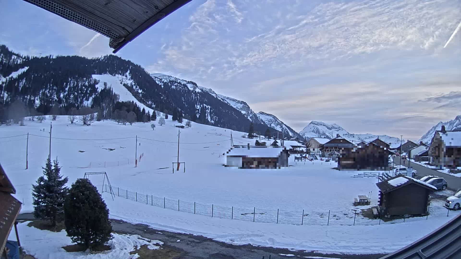 A snow-covered valley town nestled beneath mountains under a partly cloudy sky.
