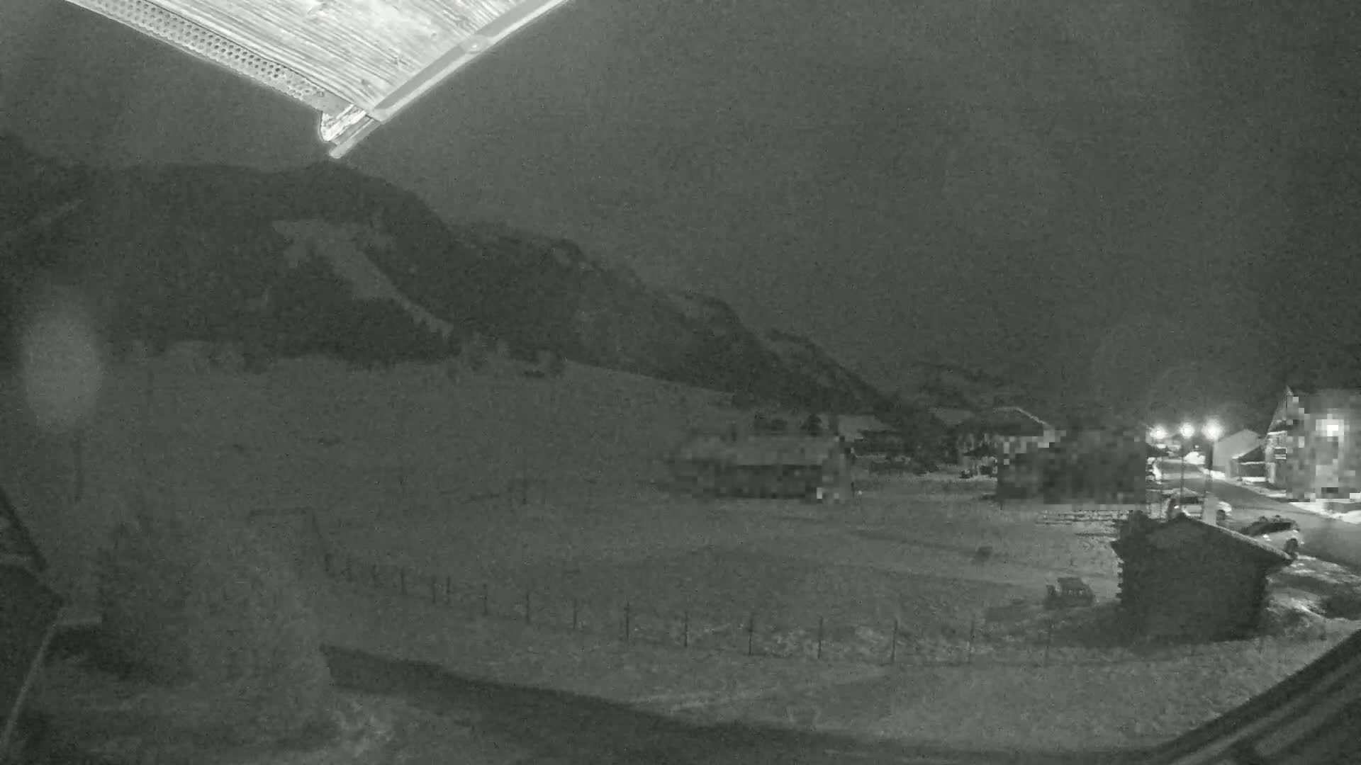 A nighttime, snow-covered landscape shows a few buildings near a mountain under a dark sky.