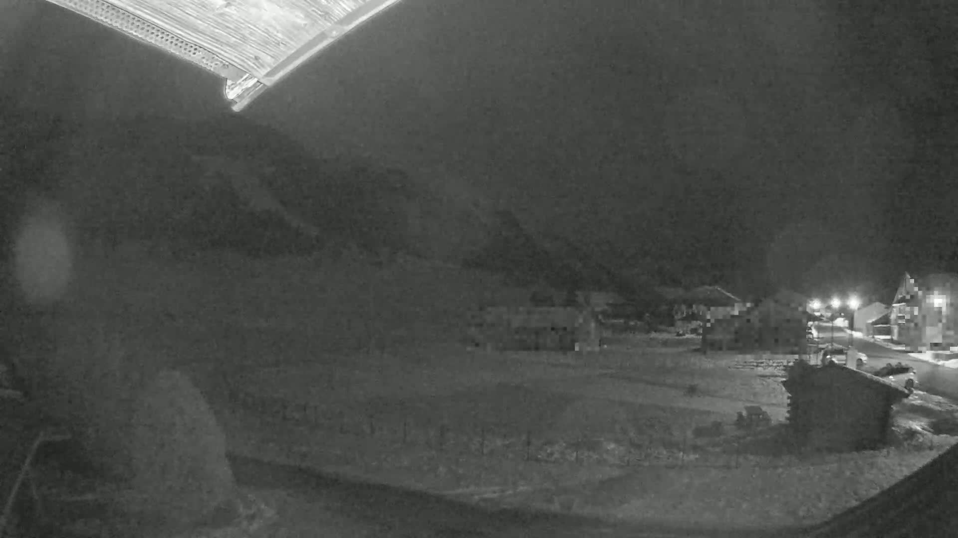 A nighttime, low-light view of a snow-covered area with several buildings and a small structure, possibly a shed, visible under a dark sky.