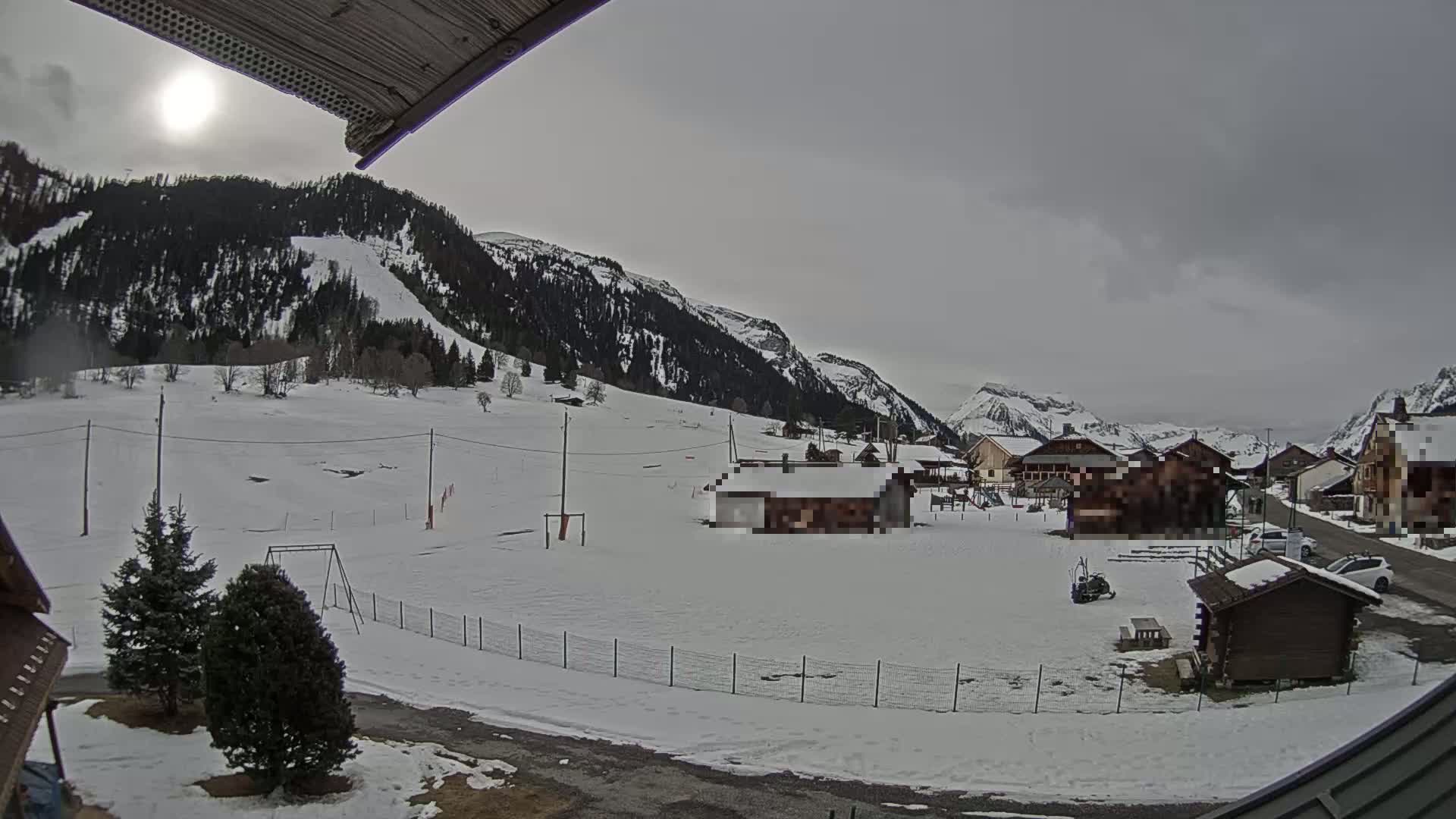 A snow-covered village nestled in a valley between mountains under a cloudy sky with a faint sun.