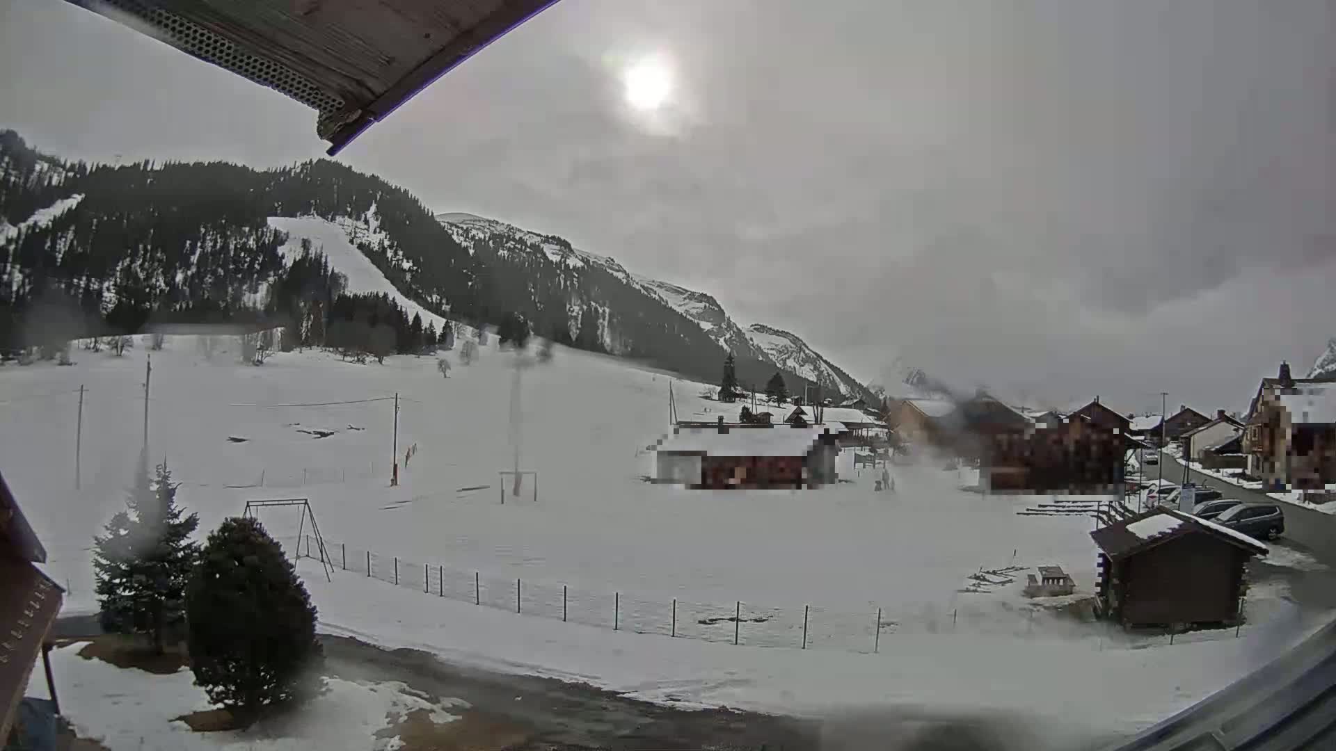 A snow-covered landscape with a few houses and a playground, nestled between mountains under an overcast sky.