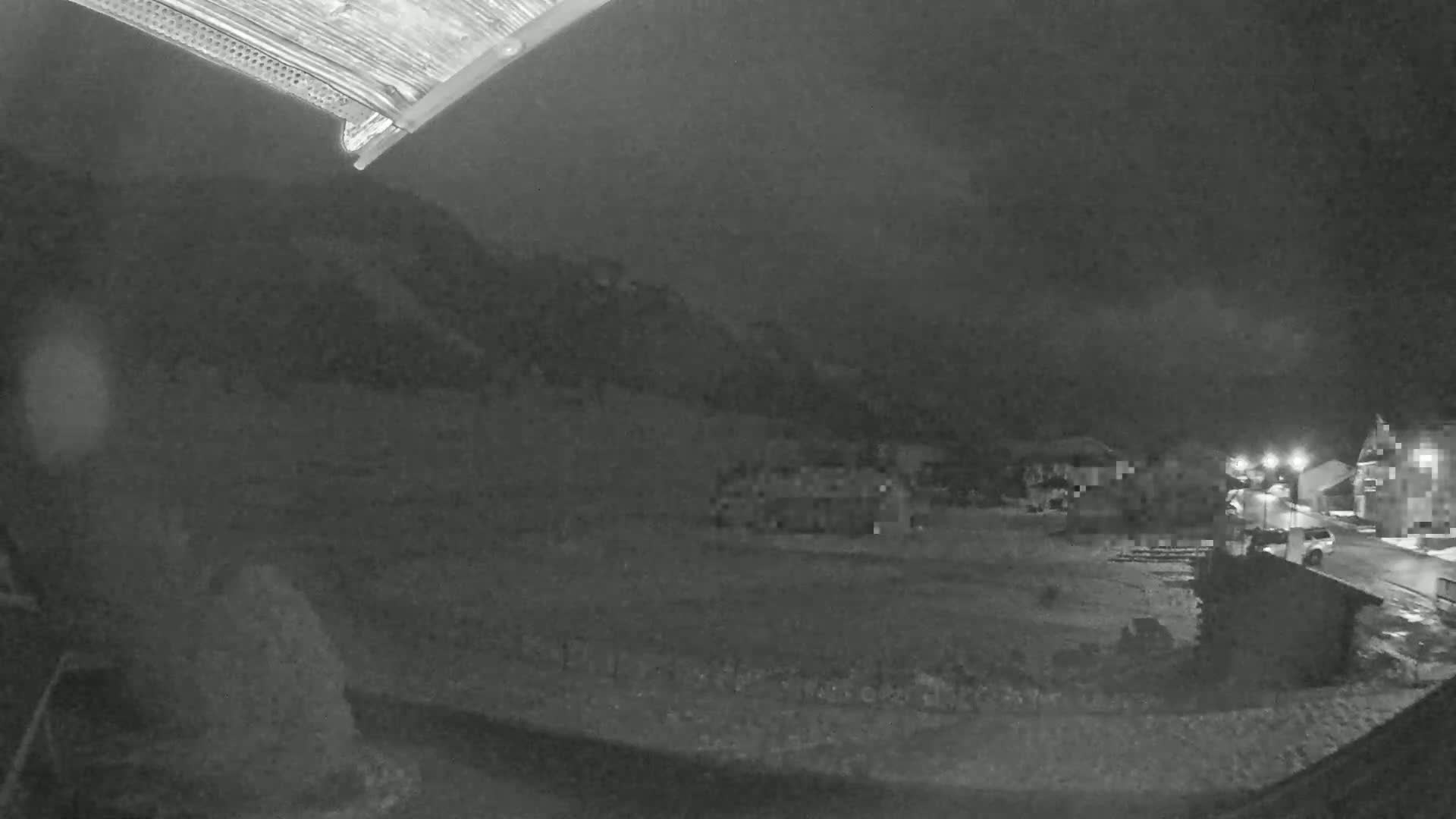 A nighttime, low-light view shows a snow-covered ground with buildings and a hill in the background under a dark, cloudy sky.
