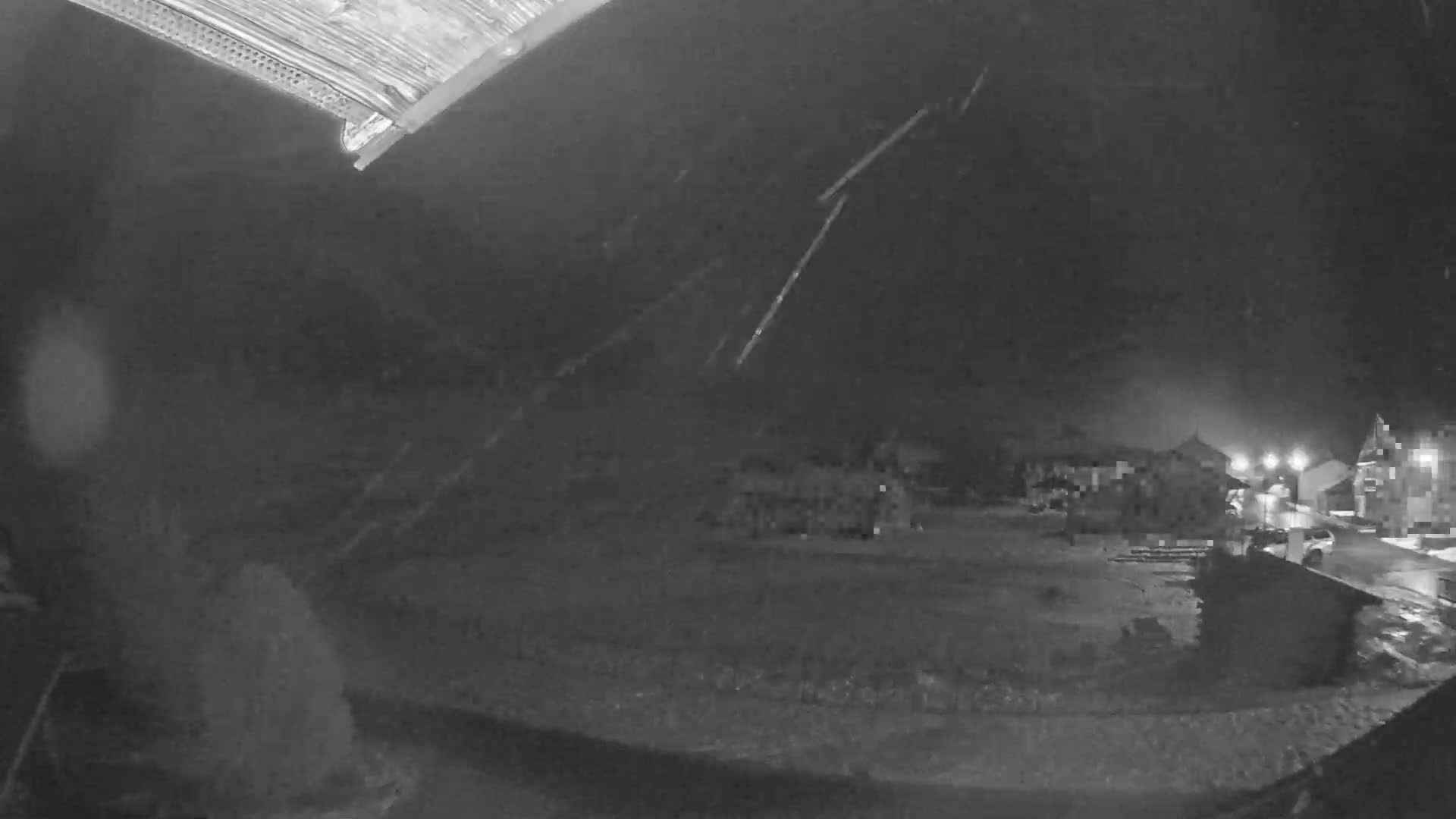 A nighttime, low-angle view shows rain falling on a dark, wet ground with some buildings faintly visible in the background.