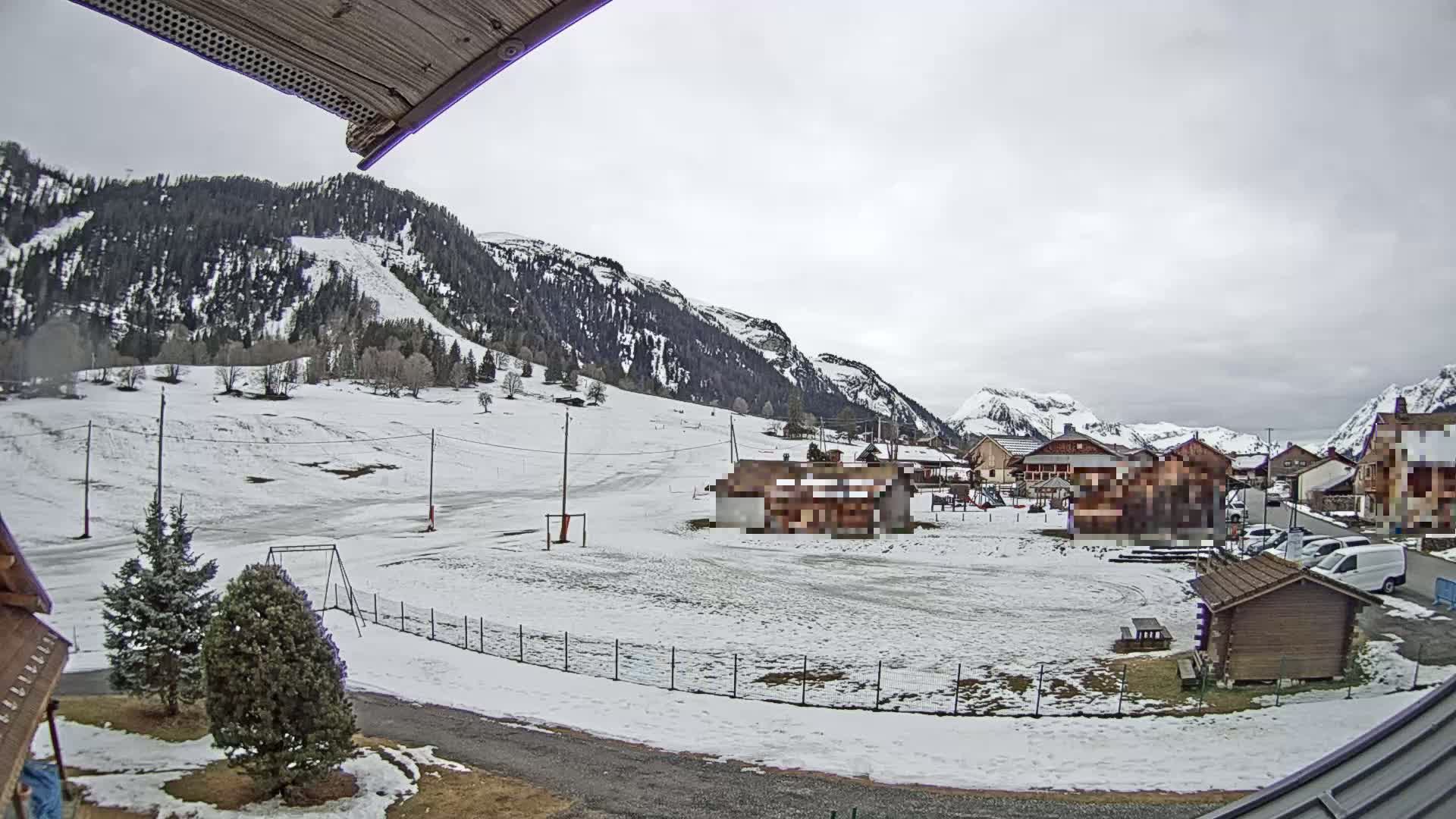 A snow-covered field with a few buildings and trees sits in front of a mountainous landscape under an overcast sky.