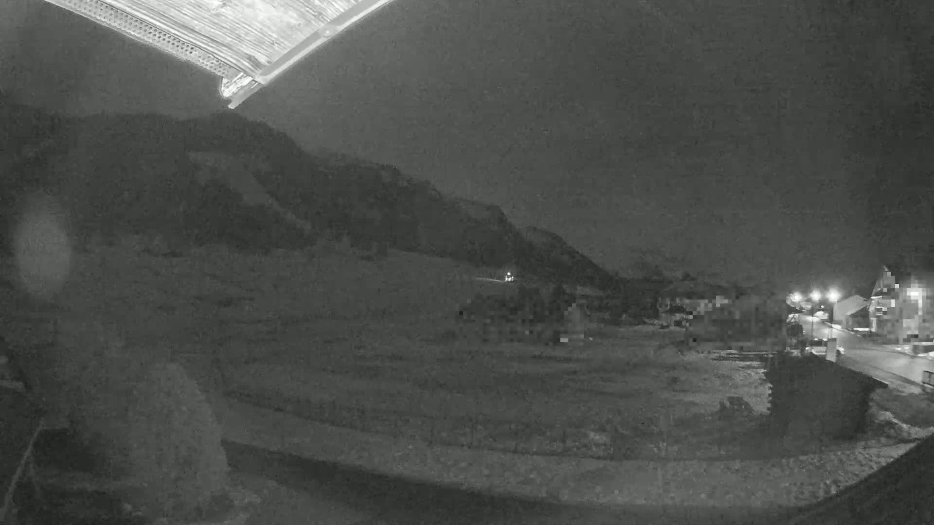 A nighttime, low-light view shows a snow-covered landscape with a few buildings and a hill in the background under a dark sky.