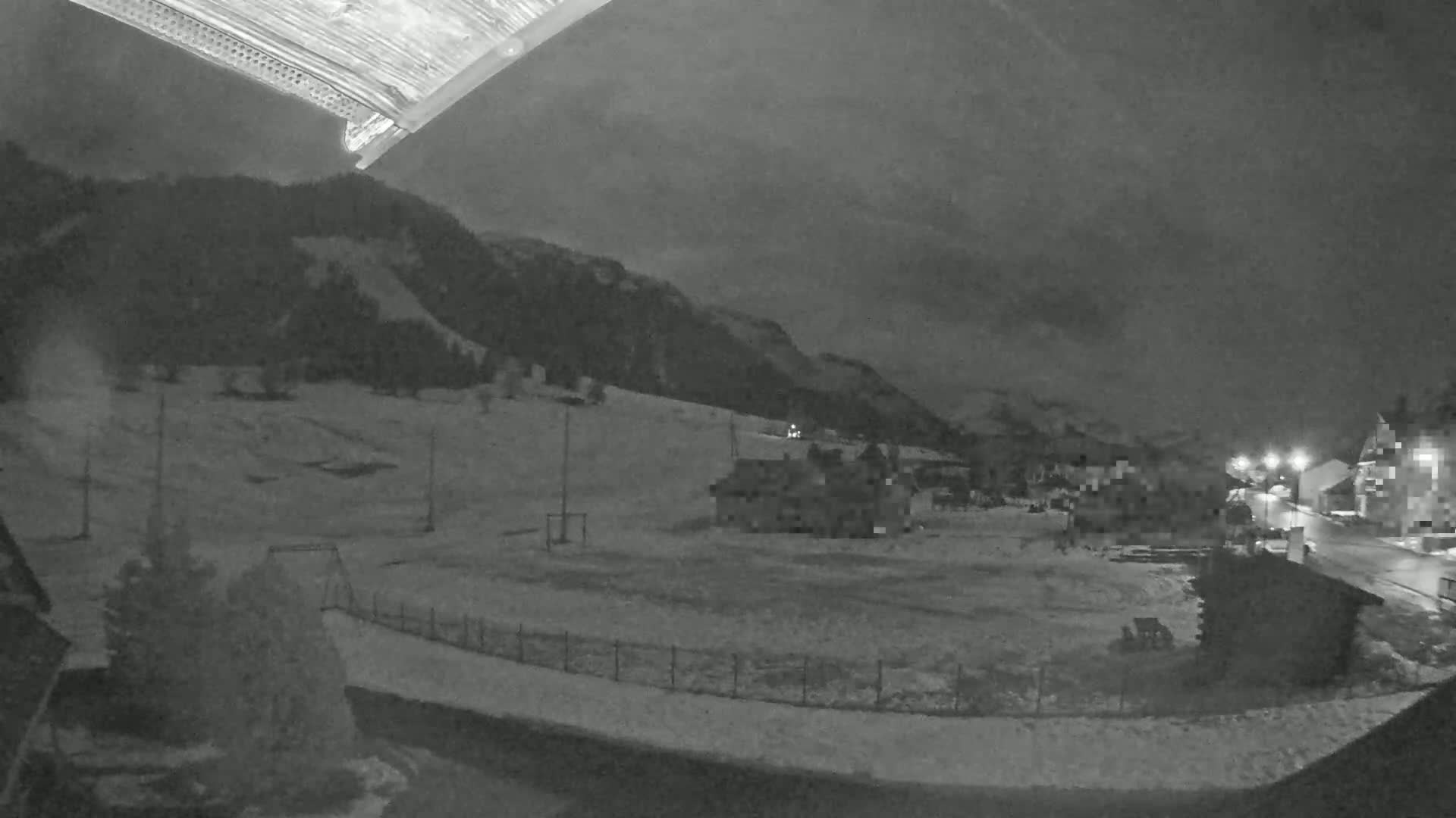A nighttime, snow-covered landscape shows a few buildings and a playground near a mountain under a dark, cloudy sky.