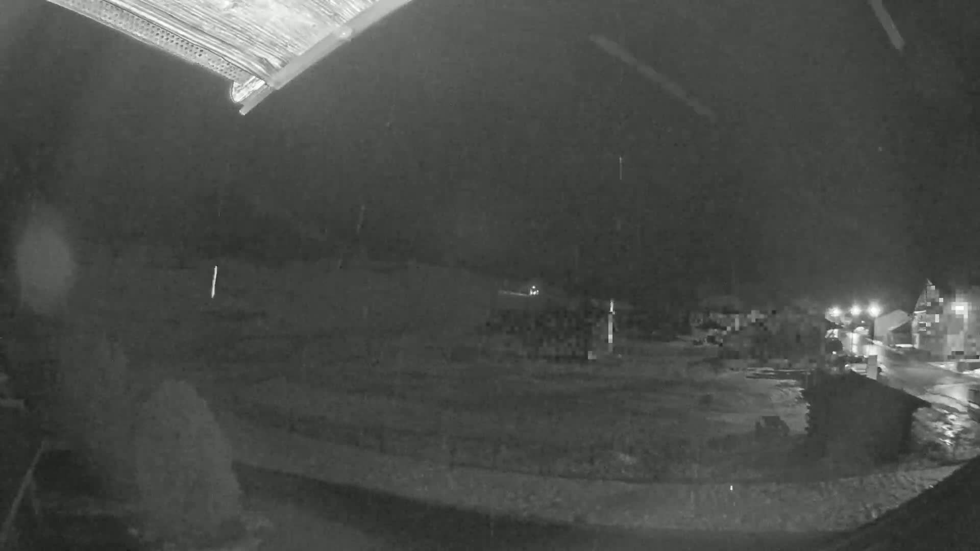 A nighttime, rainy view from under an overhang shows a dark, empty field with a few distant lights and buildings.