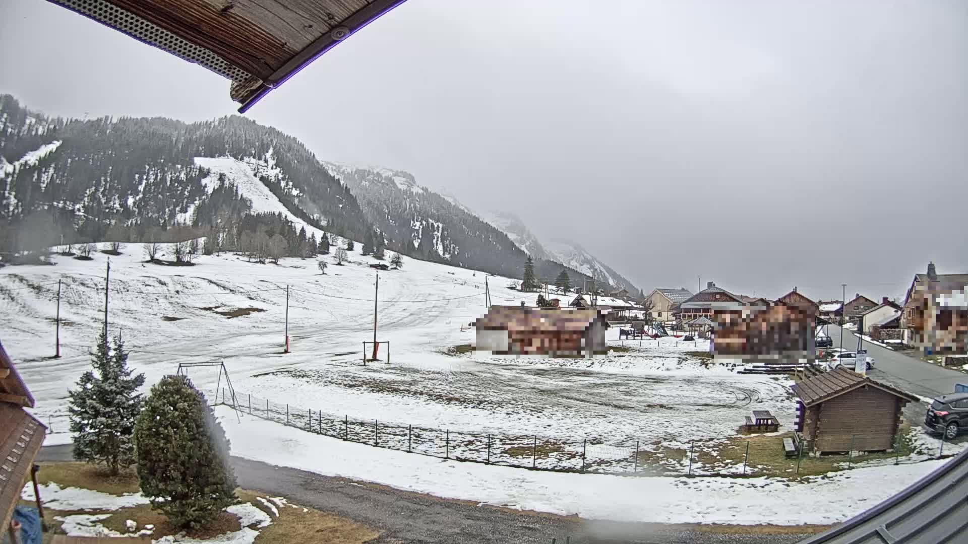 A snow-covered landscape with a small village nestled at the base of a mountain range under an overcast sky.