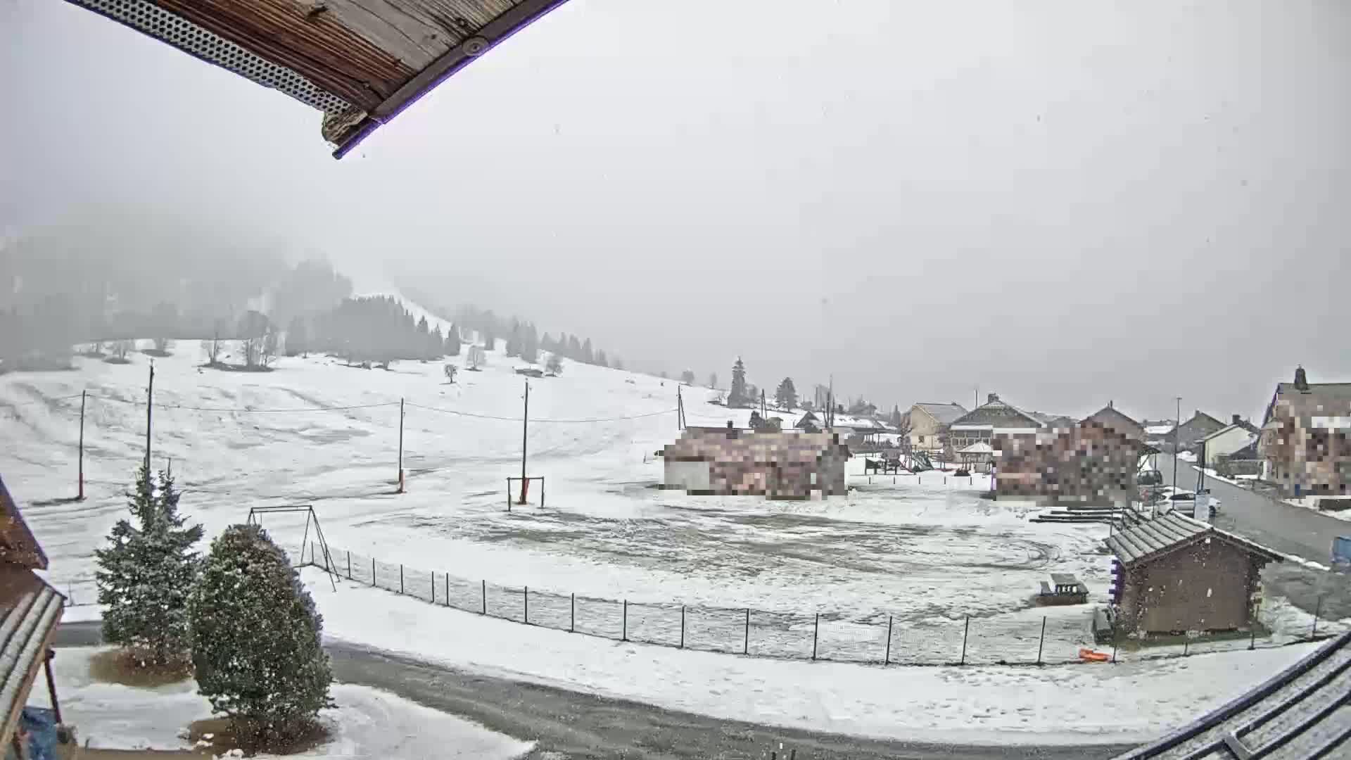 A snow-covered village in the mountains is lightly snowing, with a playground and a few buildings visible.