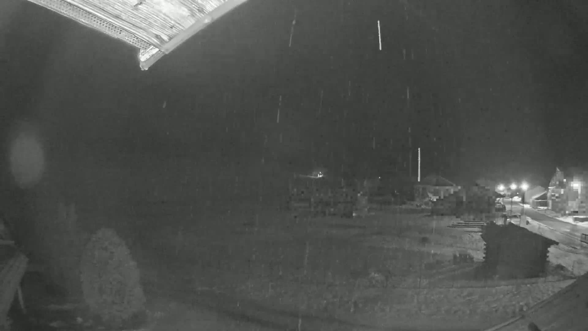 A nighttime, snowy scene shows a building in the distance and a small structure in the foreground, viewed from a slightly elevated position under a dark, overcast sky.