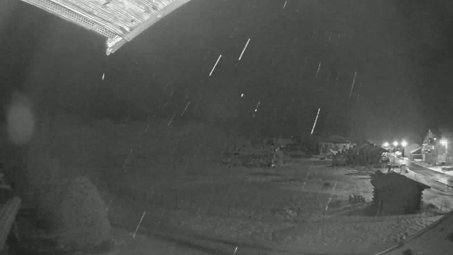 A nighttime, snowy scene shows a collection of buildings and a small structure, viewed from a slightly elevated vantage point.