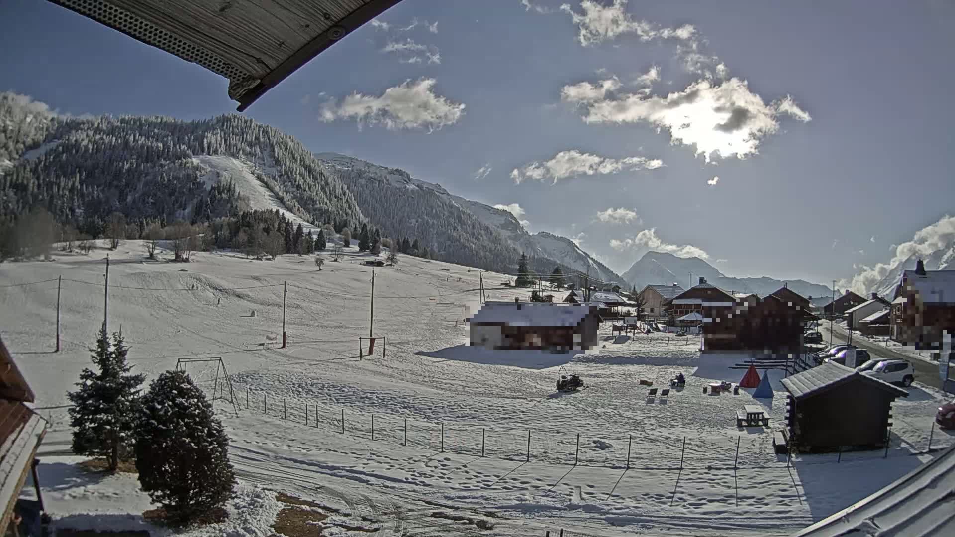 A snow-covered village nestled in a valley between snow-capped mountains under a mostly sunny sky.