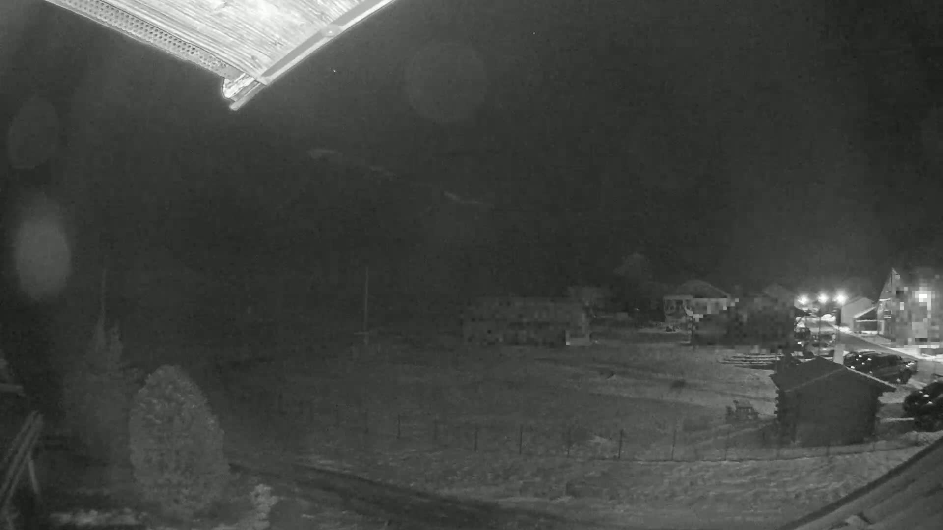 A nighttime, snow-covered scene shows a few buildings and parked cars under a dark sky.