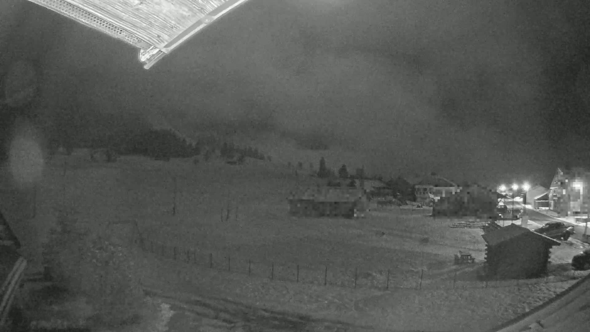 A snow-covered landscape at night, with a few buildings and some trees visible under a cloudy sky.