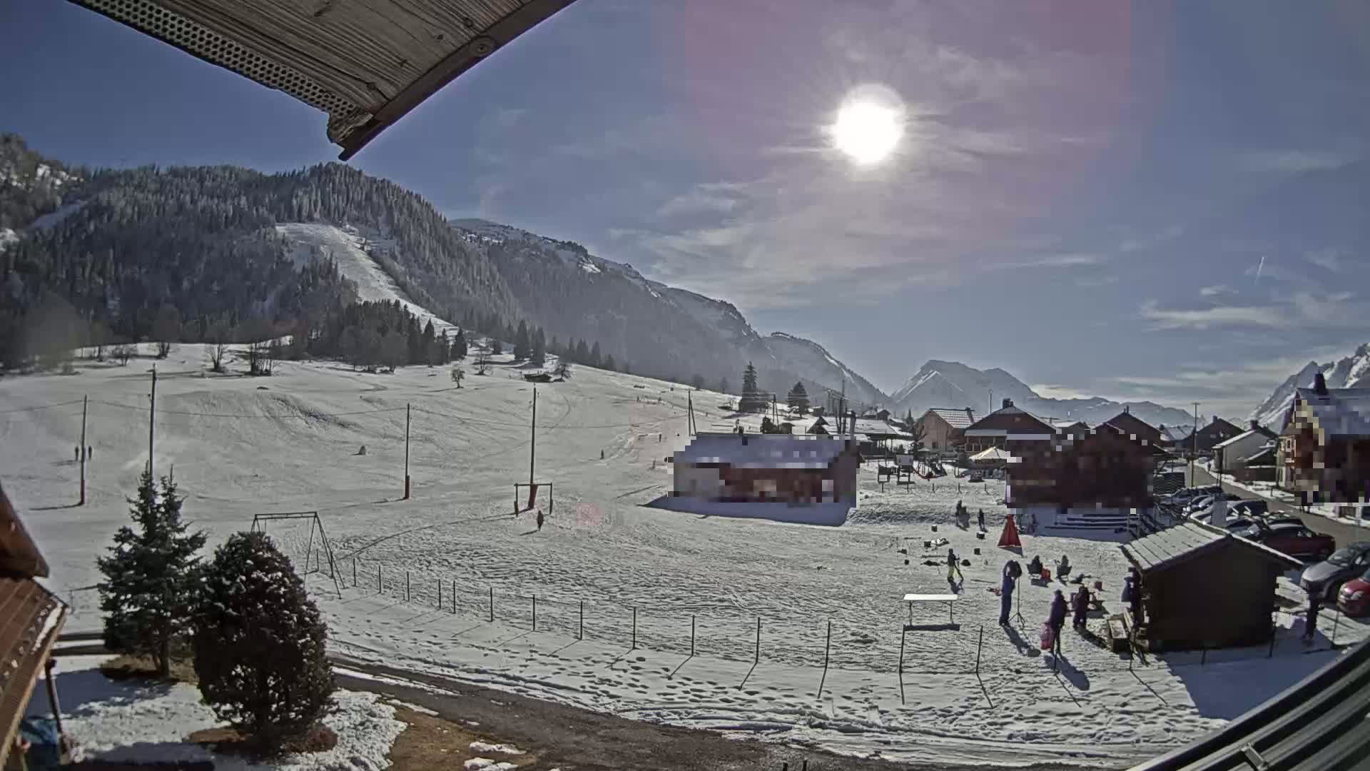 A sunny, snow-covered mountain valley scene shows a small village with people gathered near buildings and a playground, with ski slopes visible in the background.