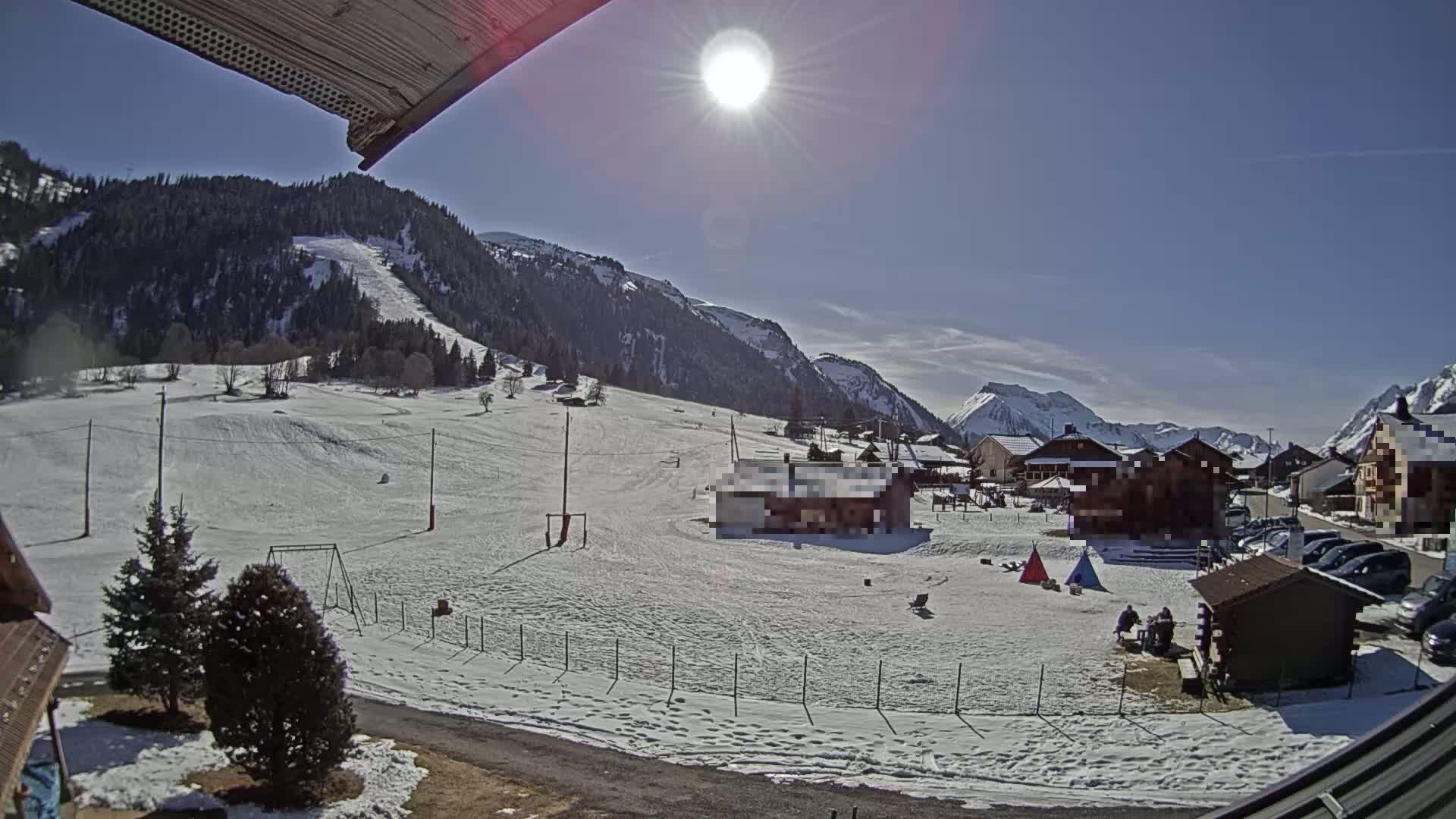 A sunny, snow-covered landscape features a small village nestled at the base of snow-capped mountains, with a playground and a few people visible in the foreground.