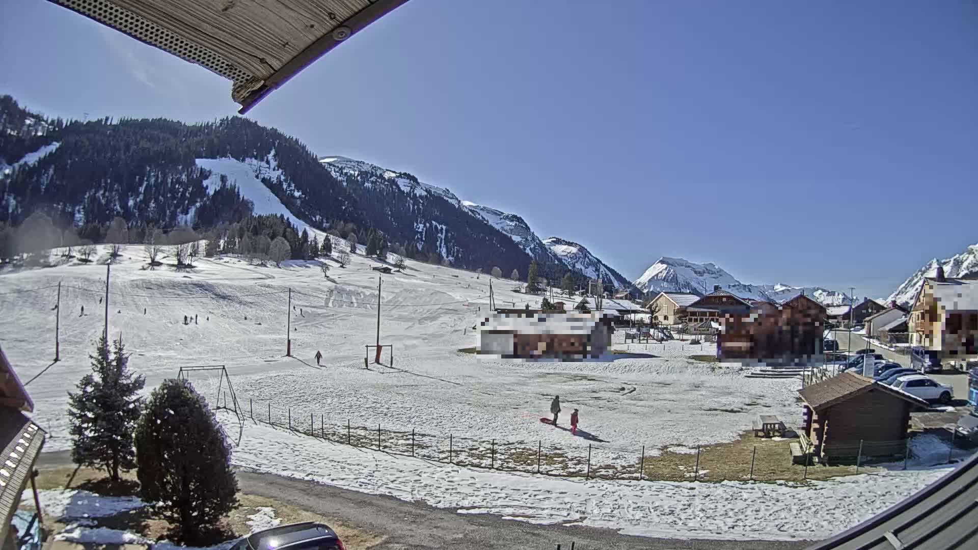 A snow-covered village nestled in a valley between mountains on a sunny day, with people enjoying winter activities on a small ski slope.