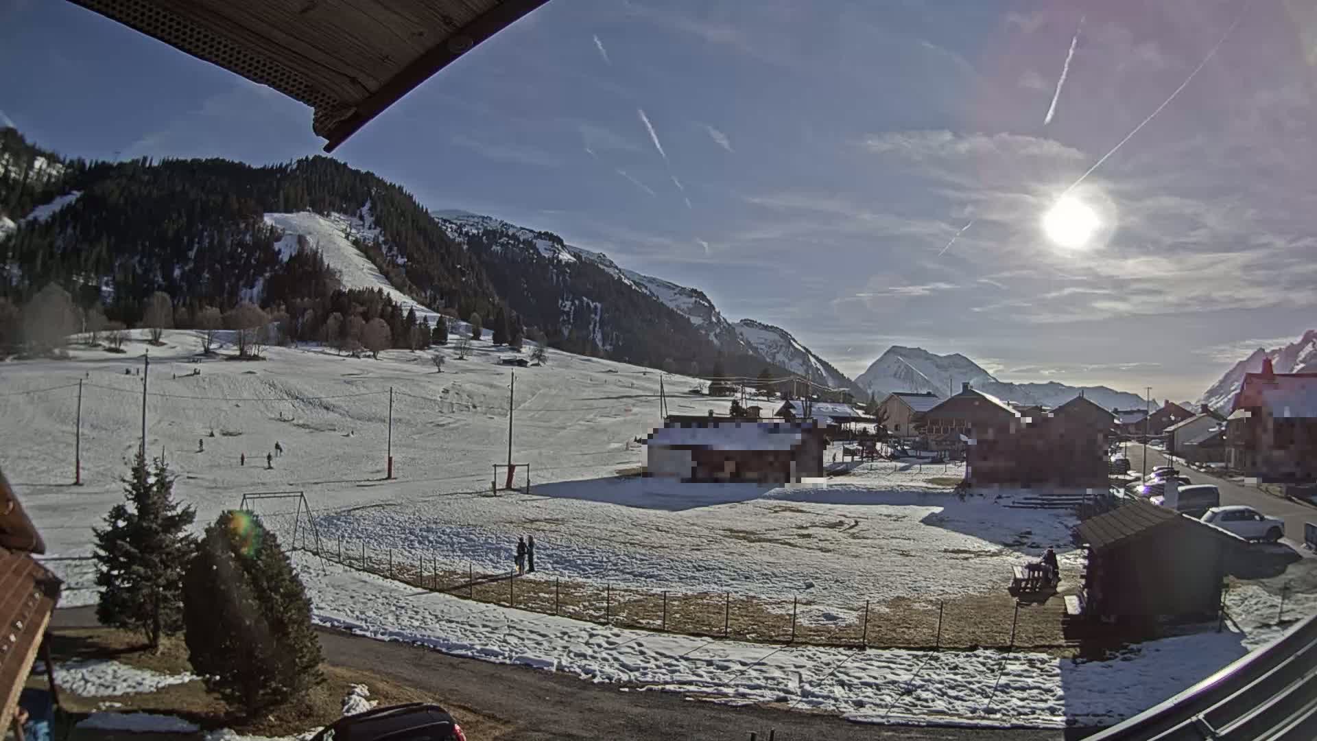 A sunny, snow-covered mountain valley scene with a small village, a few people, and a playground is visible from a slightly elevated viewpoint.