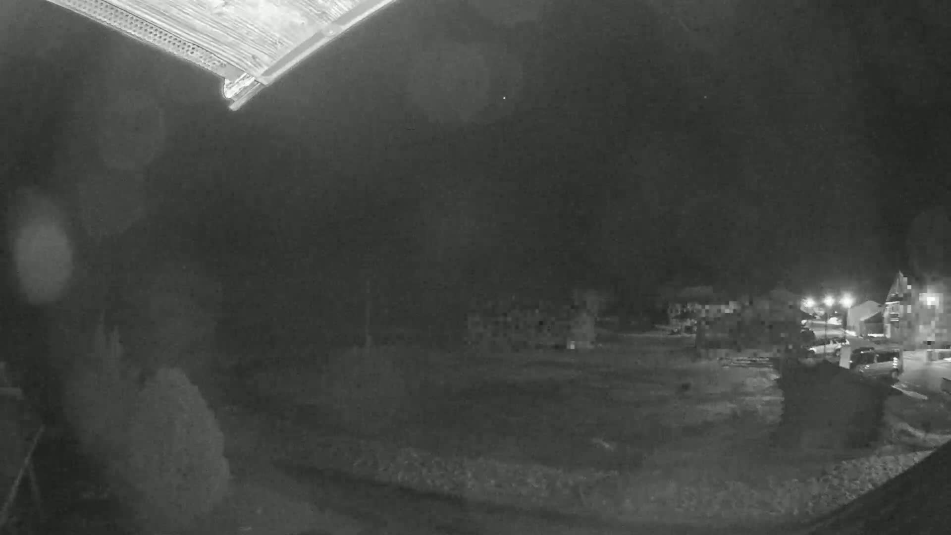 A nighttime, low-light view shows a snow-covered ground with dimly lit buildings and parked vehicles in the distance.